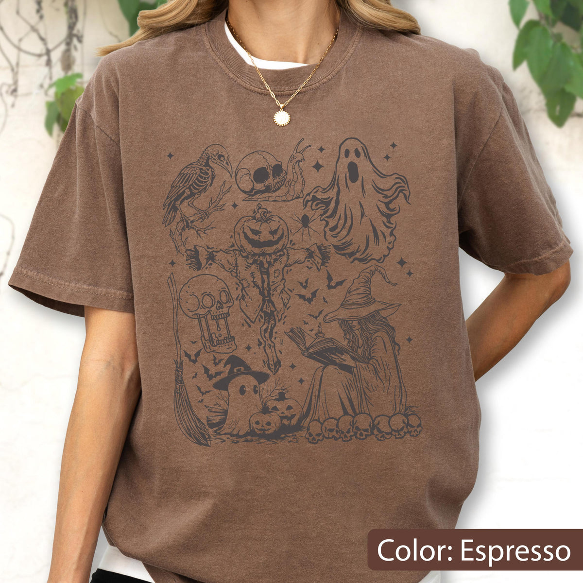 May include: Espresso-colored t-shirt featuring a Halloween-themed graphic. The design includes a crow, skulls, a scarecrow with a pumpkin head, a ghost, bats, a witch, and a broom. The shirt has a crew neck and short sleeves. The text "Color: Espresso" is at the bottom.
