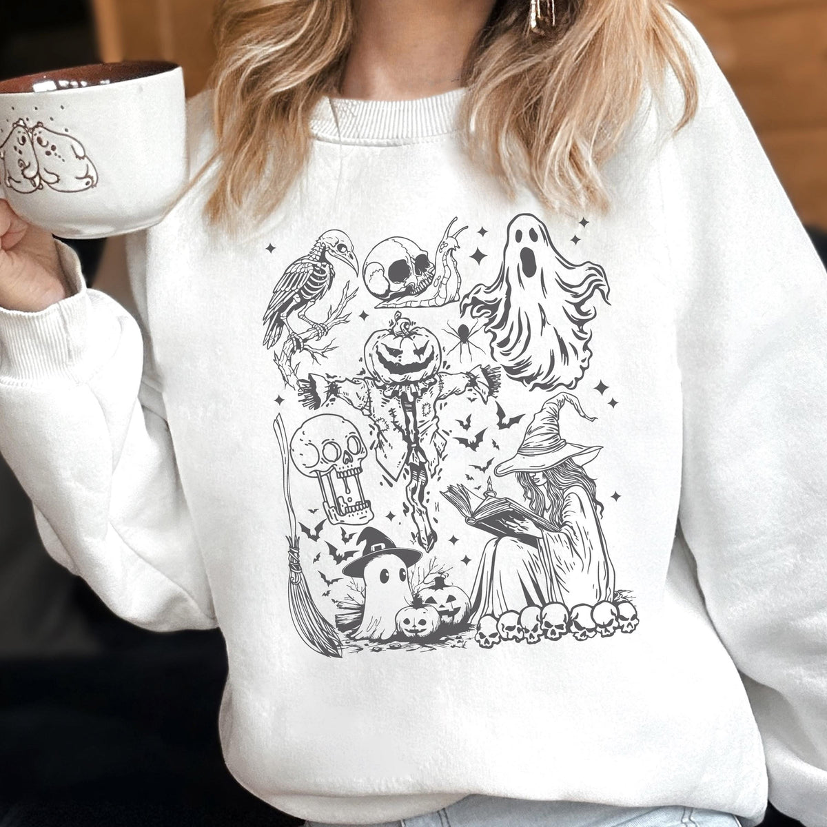May include: White sweatshirt featuring a black and white Halloween-themed graphic. The design includes a crow skeleton, skull, ghost, spider, scarecrow, bats, witch, broom, pumpkins, and skulls. A mug with a dog design is held in the image.