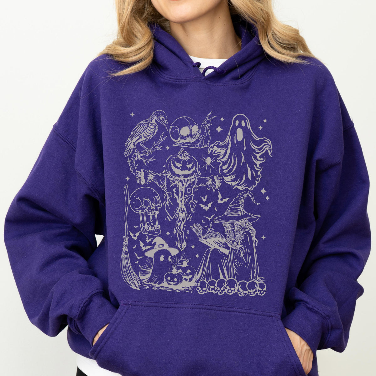 May include: A purple hoodie featuring a silver graphic of Halloween-themed illustrations. The design includes a crow, skulls, a ghost, a jack-o'-lantern, a witch, and bats. The hoodie has a hood and a front pocket.