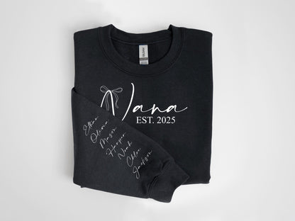 May include: Black sweatshirt with the word "Nana" in white script, a bow graphic, and the text "EST. 2025." The left sleeve displays names in white script. A cozy, casual garment.