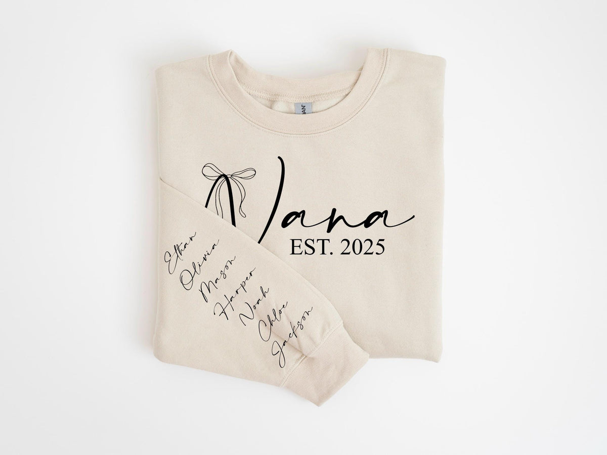 May include: A beige sweatshirt with the word "Nana" in a script font, a bow graphic, and the text "EST. 2025". The left sleeve displays names in a vertical arrangement. A cozy, personalized gift.