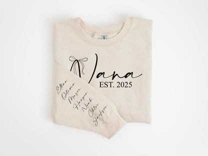 May include: A beige sweatshirt with the word "Nana" in a script font, a bow graphic, and the text "EST. 2025". The left sleeve displays names in a vertical arrangement. A cozy, personalized gift.