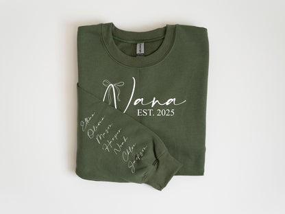May include: Olive green sweatshirt with the word "Nana" in white script, a bow graphic, and "EST. 2025". The left sleeve displays names in white script. A cozy, personalized gift.