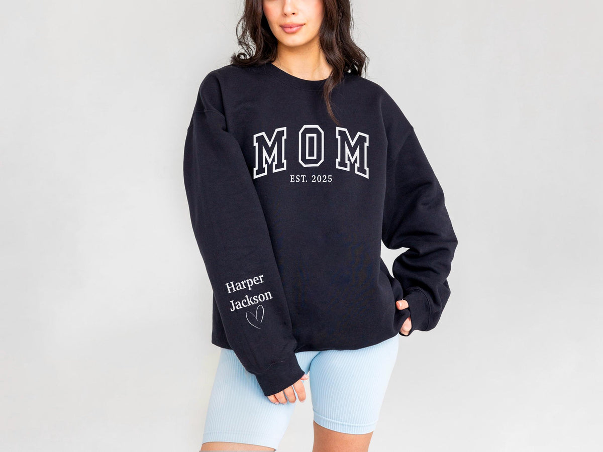 May include: A navy blue sweatshirt with the word "MOM" in white block letters, with "EST. 2025" below. The left sleeve has the name "Harper Jackson" and a heart. The sweatshirt is a casual style.