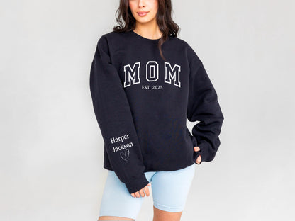 May include: A navy blue sweatshirt with the word "MOM" in white block letters, with "EST. 2025" below. The left sleeve has the name "Harper Jackson" and a heart. The sweatshirt is a casual style.