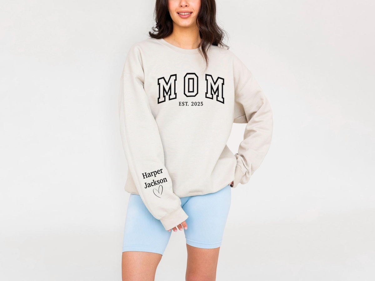 May include: A beige sweatshirt with the word "MOM" in black block letters, with "EST. 2025" below. The left sleeve has the name "Harper Jackson" and a heart. The person is wearing light blue shorts.