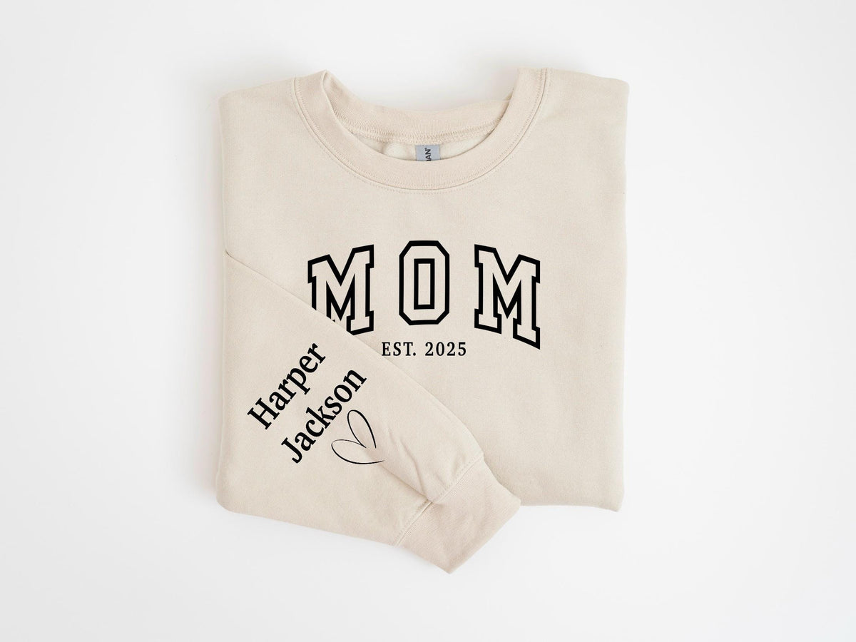 May include: A beige sweatshirt with the word "MOM" in black varsity lettering and the year "EST. 2025". The left sleeve features the name "Harper Jackson" and a heart symbol. The sweatshirt is folded neatly.