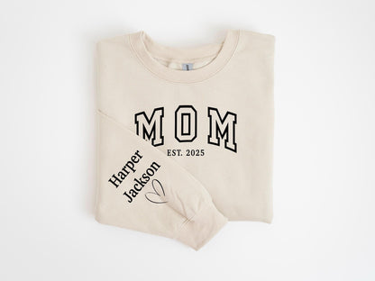 May include: A beige sweatshirt with the word "MOM" in black varsity lettering and the year "EST. 2025". The left sleeve features the name "Harper Jackson" and a heart symbol. The sweatshirt is folded neatly.