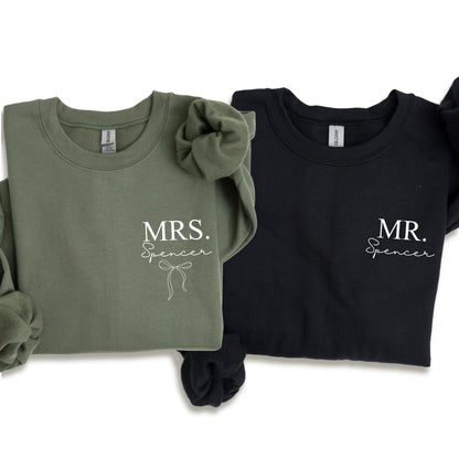 May include: Two crewneck sweatshirts, one olive green and one black. The olive green sweatshirt has the text "MRS. Spencer" with a bow graphic. The black sweatshirt has the text "MR. Spencer". Both sweatshirts are long-sleeved.