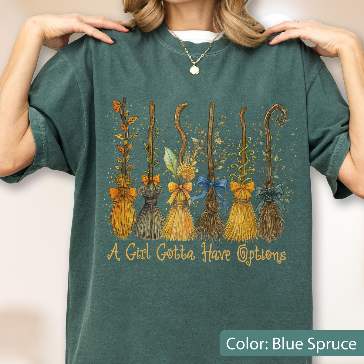 May include: A blue spruce t-shirt featuring a graphic of six distinct broomsticks decorated with leaves, flowers, and bows. The text "A Girl Gotta Have Options" is printed in a gold script font below the broomsticks on the front of the garment.