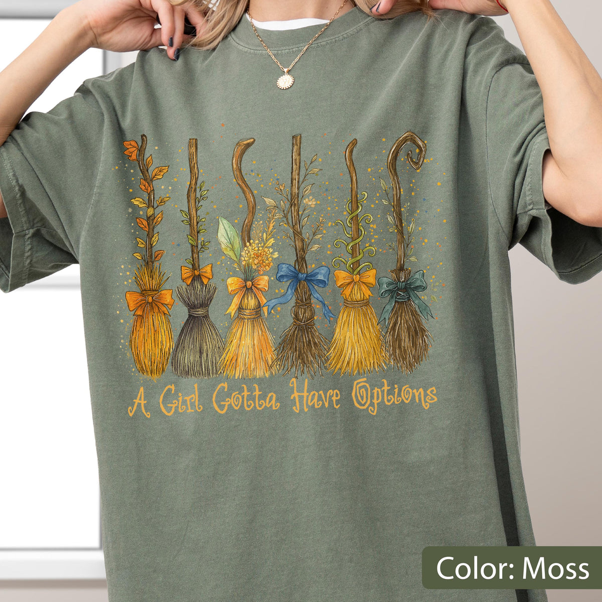 May include: A moss green t-shirt featuring a graphic print of six distinct broomsticks, each adorned with different colored bows and foliage. Below the illustration, the text reads, A Girl Gotta Have Options, in a stylized, light-colored script font.