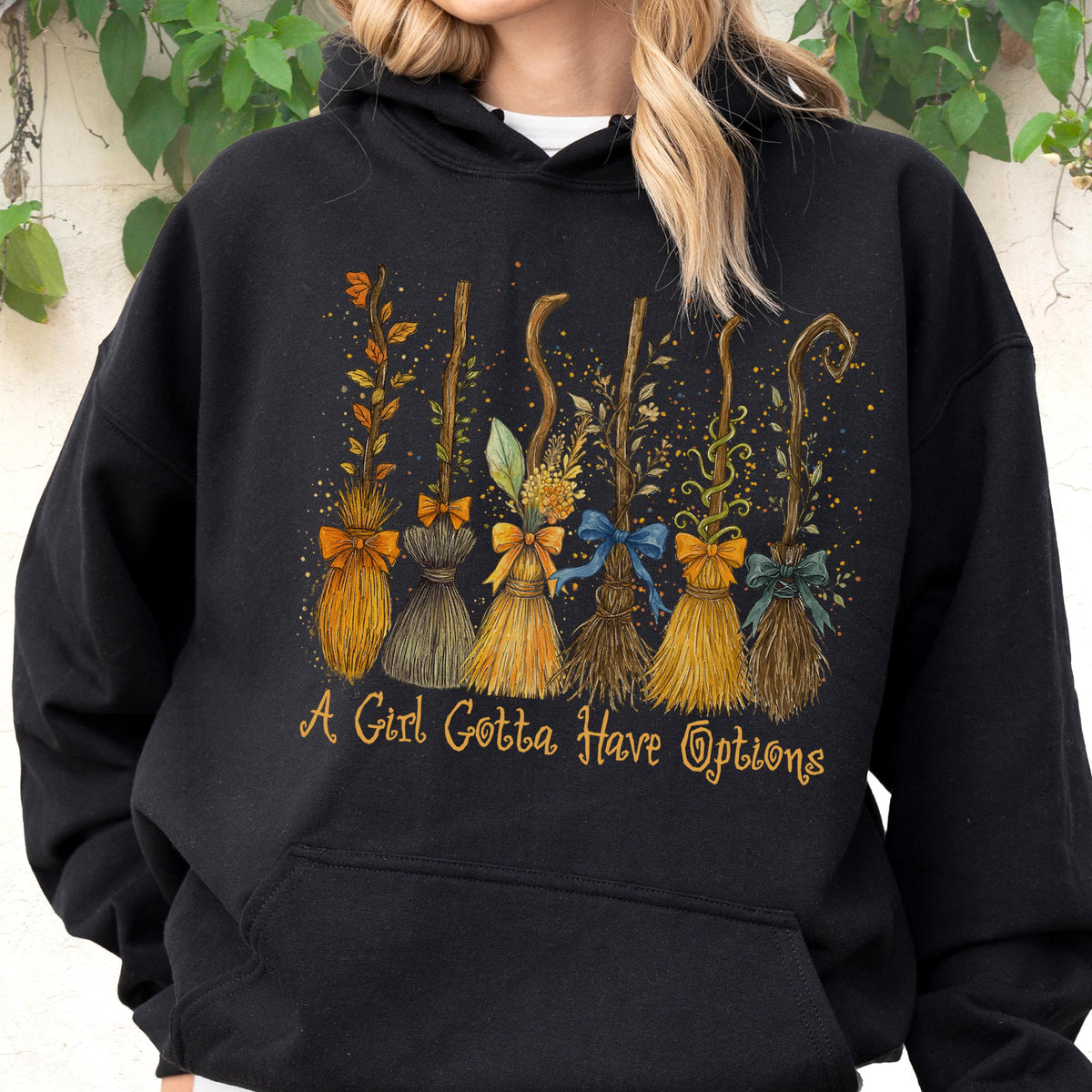 May include: A black hooded sweatshirt featuring a graphic print of six distinct broomsticks decorated with various bows and foliage. Below the broomsticks, the text reads A Girl Gotta Have Options in a stylized, golden-yellow cursive font.