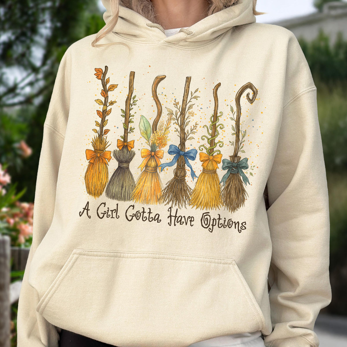 May include: A cream-colored hooded sweatshirt featuring a graphic print of six distinct broomsticks adorned with various leaves, twigs, and colorful bows. Below the illustration, the text reads, A Girl Gotta Have Options, in a dark brown script font.