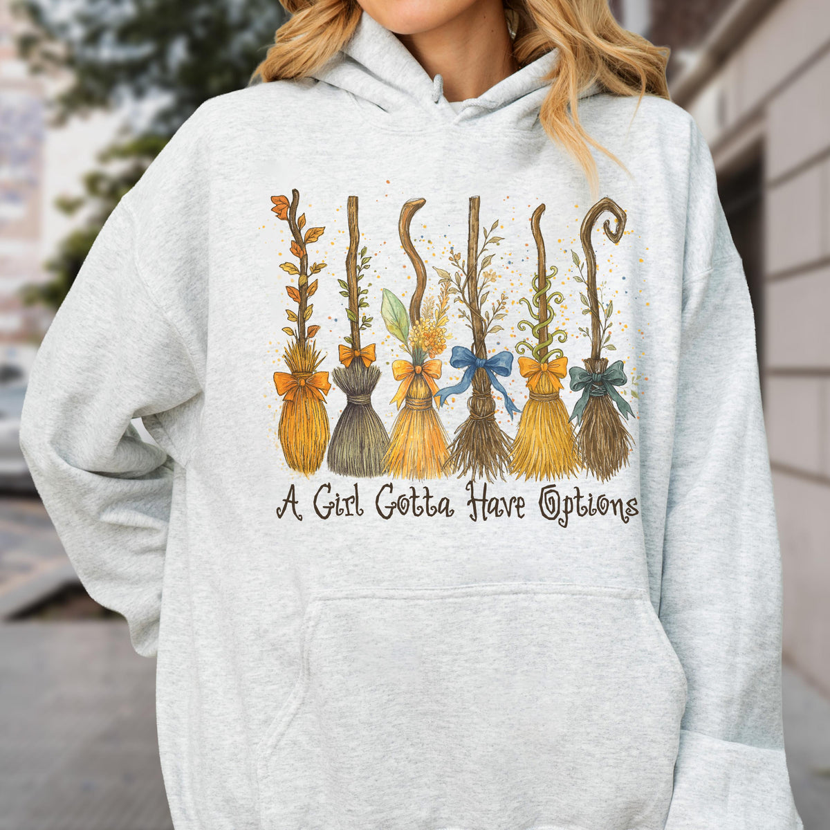May include: A light gray hooded sweatshirt featuring a graphic print of six distinct broomsticks decorated with bows and foliage. Below the illustration, the text reads, A Girl Gotta Have Options, in a dark, stylized, cursive-inspired font.