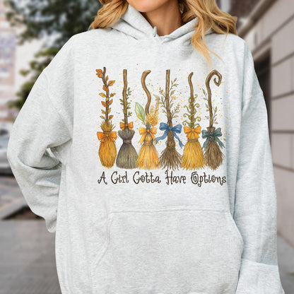 May include: A light gray hooded sweatshirt featuring a graphic print of six distinct broomsticks decorated with bows and foliage. Below the illustration, the text reads, A Girl Gotta Have Options, in a dark, stylized, cursive-inspired font.
