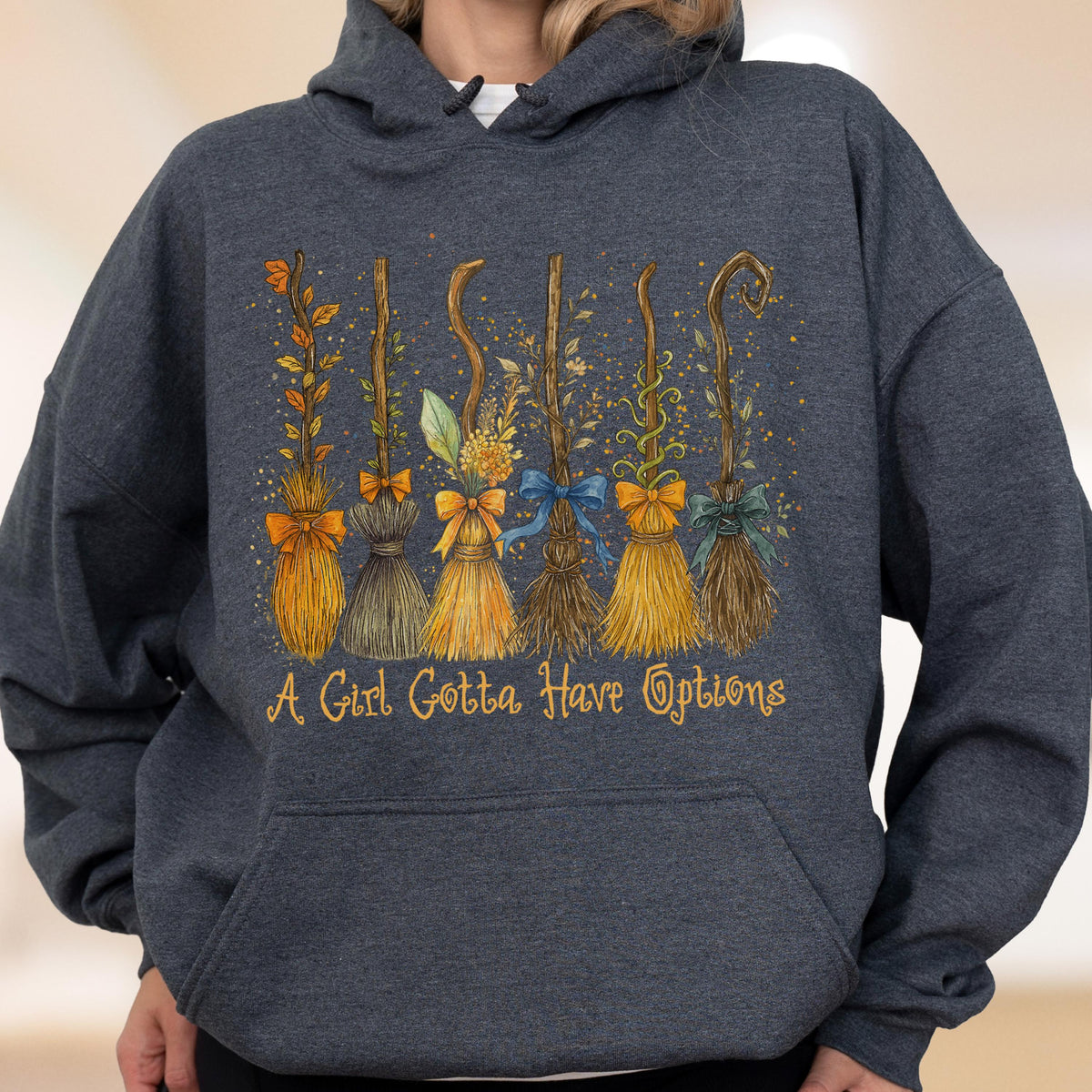 May include: A dark heather gray hooded sweatshirt featuring a graphic of six distinct broomsticks decorated with colorful bows and autumn foliage. Below the brooms, the text reads, A Girl Gotta Have Options, in a stylized, golden-yellow script font.