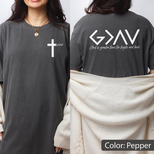 May include: A pepper-colored t-shirt featuring a white cross graphic on the front left chest with the text G>AV. The back displays the text G>AV and the phrase God is greater than the highs and lows in white lettering against the dark fabric.