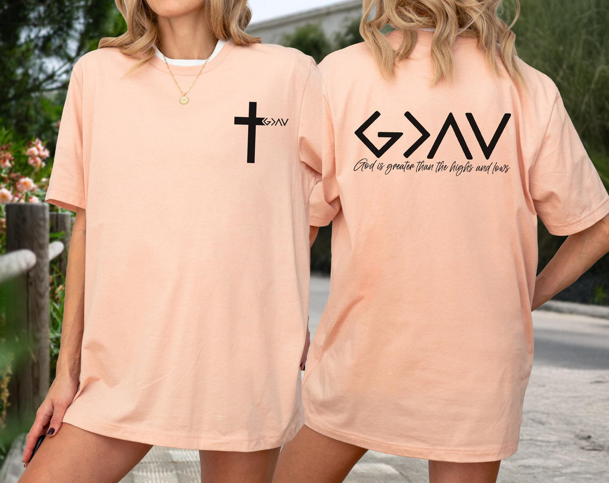 May include: Peach-colored t-shirt with a black cross and the letters "G>AV" on the front. The back of the shirt features the same letters and the phrase "God is greater than the highs and fears" in black.