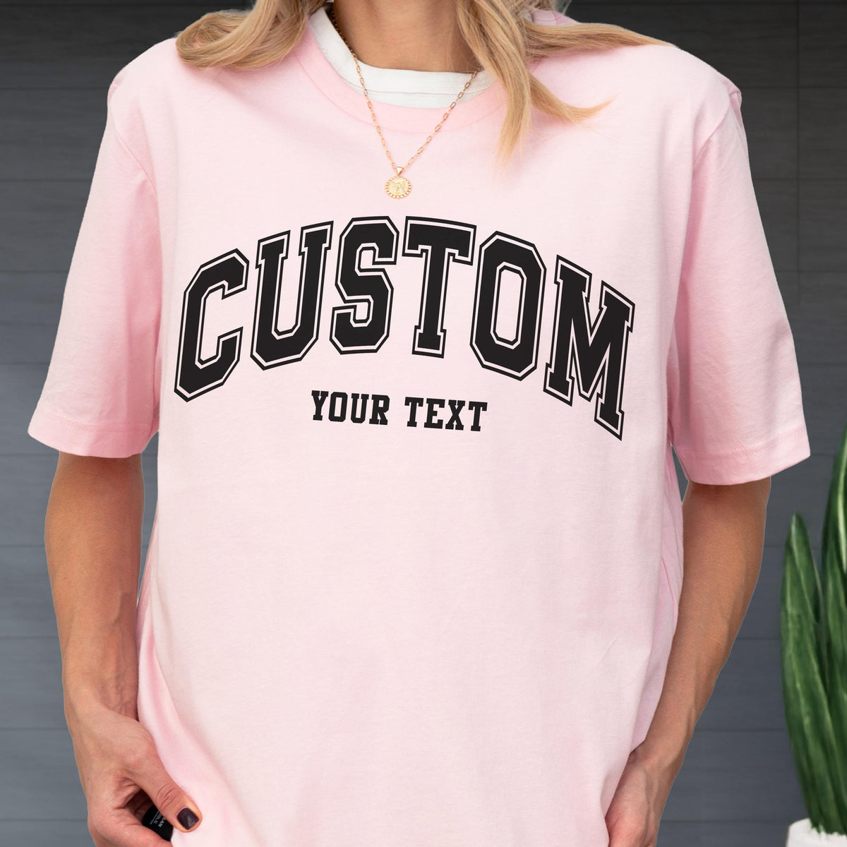 May include: A light pink t-shirt with the word "CUSTOM" arched across the chest in black block letters with a white outline. Below the word "CUSTOM" is the text "YOUR TEXT" in a smaller, black font.