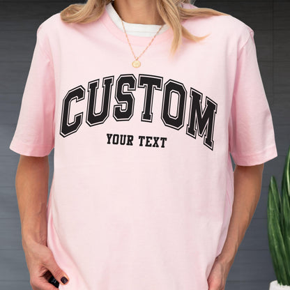 May include: A light pink t-shirt with the word "CUSTOM" arched across the chest in black block letters with a white outline. Below the word "CUSTOM" is the text "YOUR TEXT" in a smaller, black font.