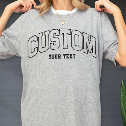 May include: A light gray t-shirt with the word "CUSTOM" in a black, collegiate-style font across the chest. Below the word "CUSTOM" is the text "YOUR TEXT" in a smaller, black font. The shirt has a crew neck and short sleeves.