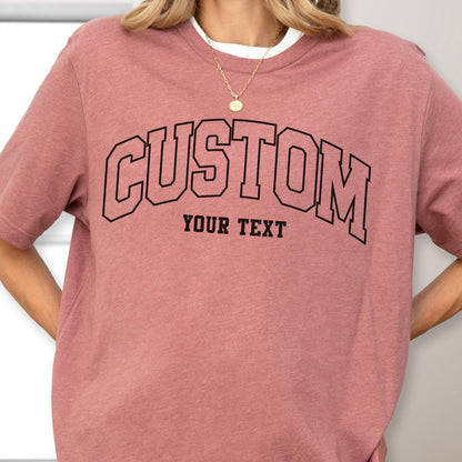 May include: A dusty rose-colored t-shirt with the word "CUSTOM" arched across the chest in a black, collegiate-style font. Below the word "CUSTOM" is the text "YOUR TEXT" in a smaller, matching font. The shirt has a crew neck and short sleeves.