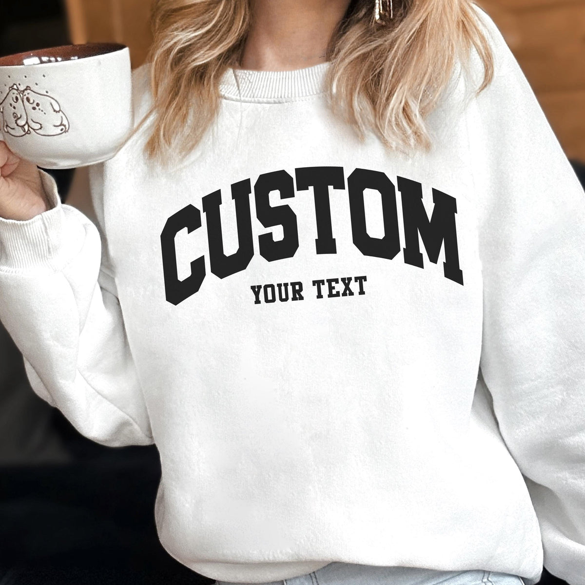 May include: White sweatshirt with the word "CUSTOM" in large, black, block letters across the chest, with "YOUR TEXT" below. The sweatshirt is a crew neck style. A white mug with a dog illustration is held in the image.