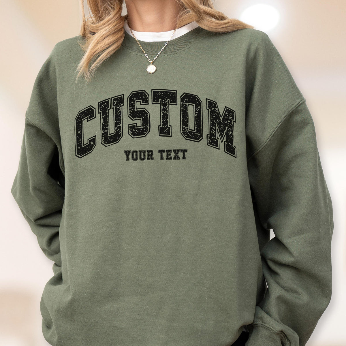 May include: An olive green sweatshirt with the word "CUSTOM" in a black, collegiate-style font across the chest. Below the word "CUSTOM" is the text "YOUR TEXT" in a smaller font. The sweatshirt has long sleeves and a crew neck.