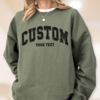 May include: An olive green sweatshirt with the word "CUSTOM" in a black, collegiate-style font across the chest. Below the word "CUSTOM" is the text "YOUR TEXT" in a smaller font. The sweatshirt has long sleeves and a crew neck.