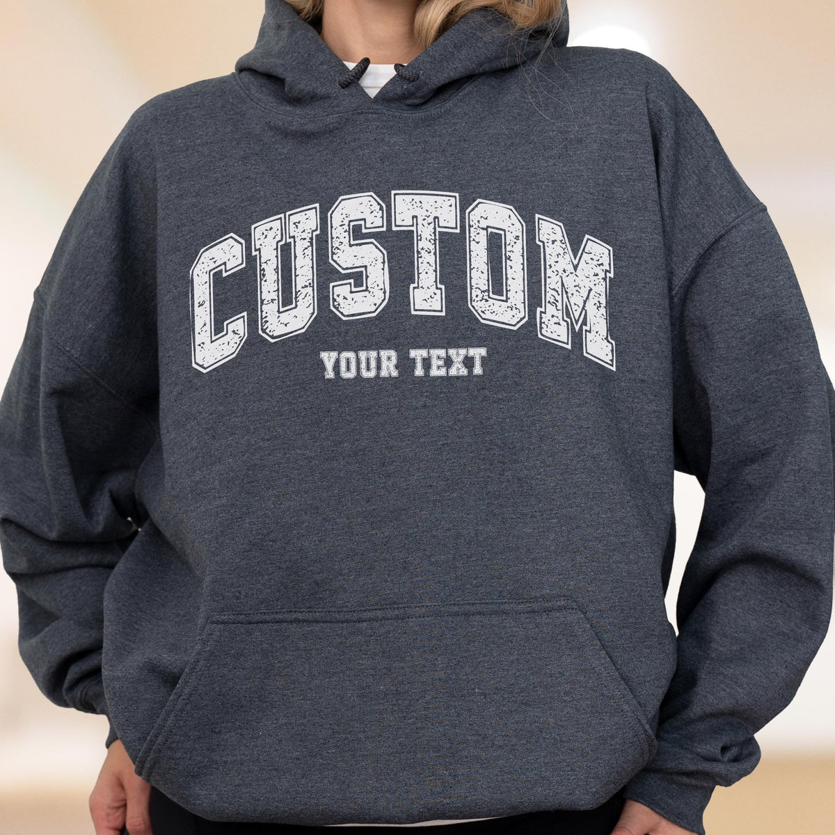 May include: A dark gray hooded sweatshirt with a white distressed "CUSTOM" arched text above the words "YOUR TEXT". The sweatshirt has a front pocket and a drawstring hood. The overall style is casual and comfortable.