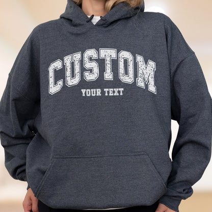 May include: A dark gray hooded sweatshirt with a white distressed "CUSTOM" arched text above the words "YOUR TEXT". The sweatshirt has a front pocket and a drawstring hood. The overall style is casual and comfortable.