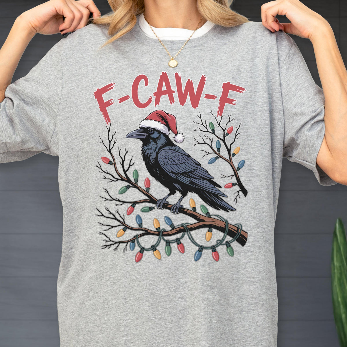 May include: A heather gray t-shirt featuring a black raven wearing a red and white Santa hat perched on a bare tree branch. The branch is decorated with colorful Christmas lights. Above the raven, the text F-CAW-F is printed in red letters.