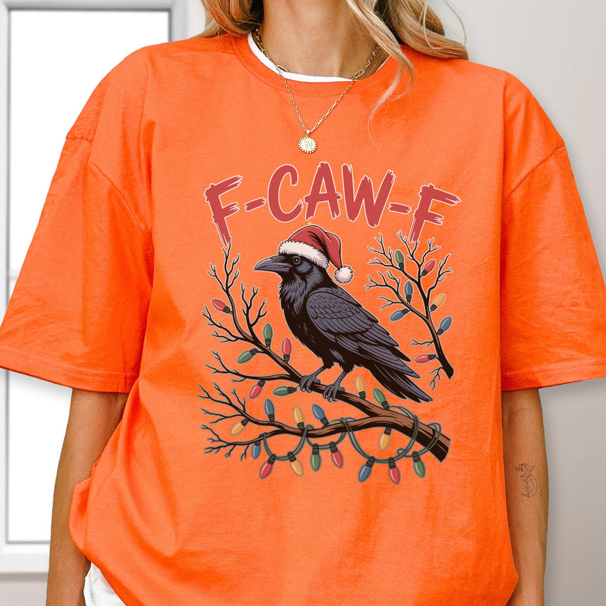 May include: A bright orange t-shirt featuring a graphic of a black crow wearing a red and white Santa hat, perched on a tree branch decorated with colorful Christmas lights. The text F-CAW-F is printed in red above the crow illustration.