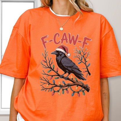 May include: A bright orange t-shirt featuring a graphic of a black crow wearing a red and white Santa hat, perched on a tree branch decorated with colorful Christmas lights. The text F-CAW-F is printed in red above the crow illustration.