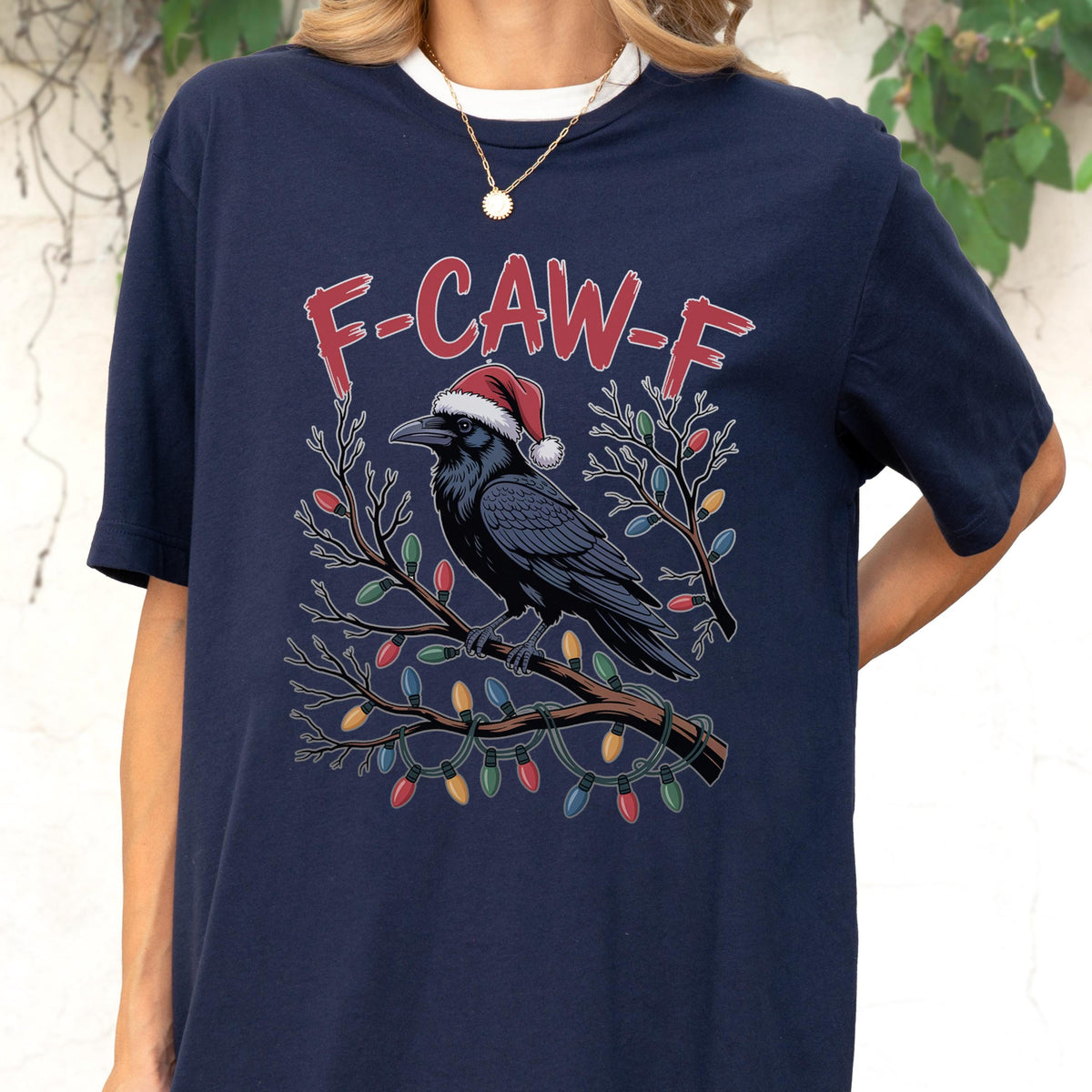 May include: A navy blue t-shirt featuring a graphic of a black crow wearing a red and white Santa hat, perched on a brown tree branch wrapped in colorful Christmas lights. The text F-CAW-F is printed in red above the crow in a distressed font.