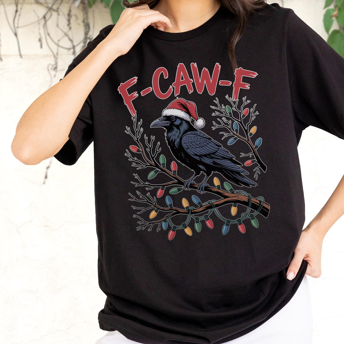 May include: A black short-sleeve t-shirt featuring a graphic of a black crow wearing a red and white Santa hat, perched on a bare tree branch wrapped in colorful Christmas lights, with the red text F-CAW-F printed above the bird illustration.
