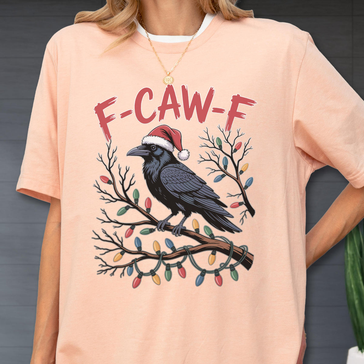 May include: A peach-colored t-shirt featuring a graphic of a black raven wearing a red and white Santa hat, perched on a bare tree branch wrapped in colorful Christmas lights. The text F-CAW-F is printed in red above the raven illustration.