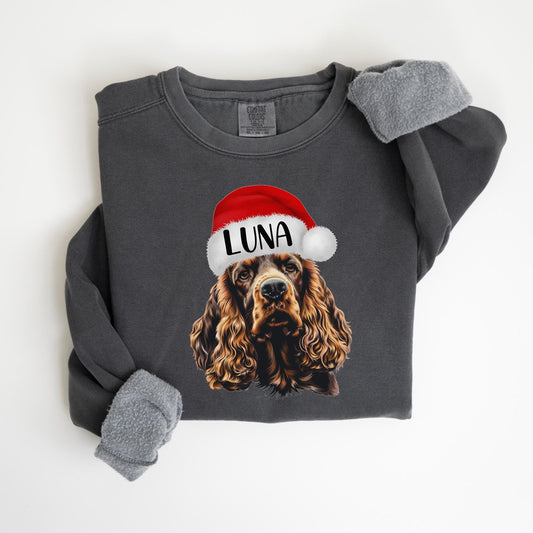 May include: Dark gray sweatshirt featuring a Cocker Spaniel wearing a Santa hat with the name "LUNA" printed on it. The sleeves have a gray, fuzzy lining. The sweatshirt is a comfortable, casual style.