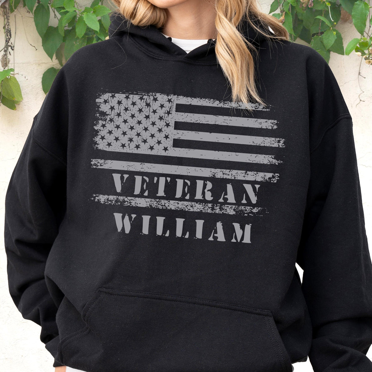 May include: Black hooded sweatshirt featuring a distressed gray American flag design. The text "VETERAN WILLIAM" is printed below the flag. The sweatshirt has a front pocket and a drawstring hood.