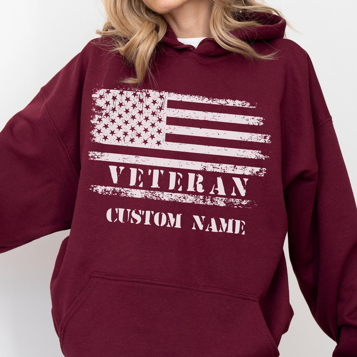 May include: Burgundy hoodie featuring a distressed American flag design in white. The word "VETERAN" is printed below the flag, with space for a custom name. The hoodie has a drawstring hood and a front pocket.