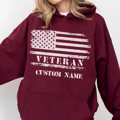 May include: Burgundy hoodie featuring a distressed American flag design in white. The word "VETERAN" is printed below the flag, with space for a custom name. The hoodie has a drawstring hood and a front pocket.