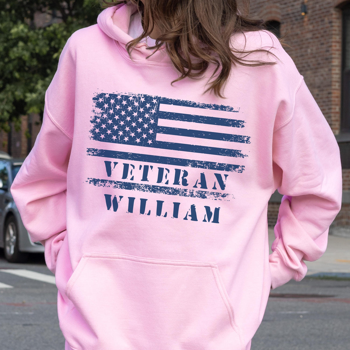 May include: A light pink hoodie featuring a distressed blue American flag design on the back. Below the flag, the word "VETERAN" is printed, followed by the name "WILLIAM". The hoodie has a front pocket and a hood.