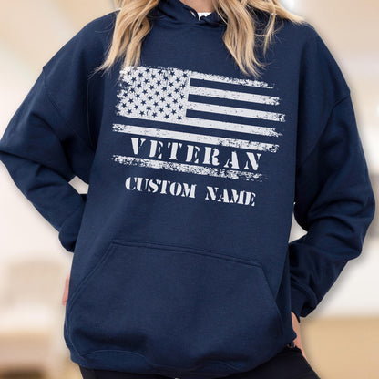 May include: Navy blue hooded sweatshirt featuring a distressed American flag design in white. The word "VETERAN" is printed across the center, with "CUSTOM NAME" below. The sweatshirt has a front pocket and a drawstring hood.