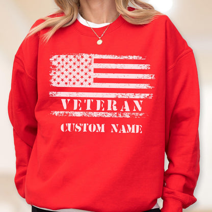 May include: A red sweatshirt featuring a distressed American flag design in white. The word "VETERAN" is printed below the flag, with space for a custom name. The sweatshirt is a comfortable, casual style.