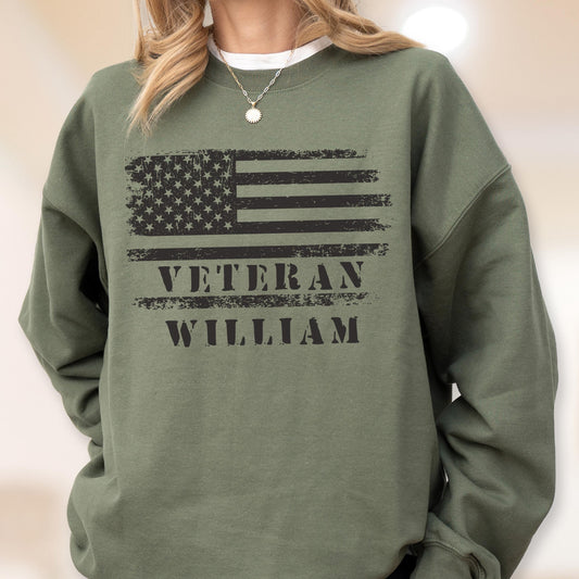 May include: An olive green sweatshirt featuring a distressed American flag design in black. The text "VETERAN WILLIAM" is printed below the flag. The sweatshirt has long sleeves and a crew neck.