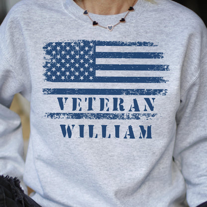 May include: A light gray sweatshirt featuring a distressed blue American flag design. Below the flag, the word "VETERAN" is printed above the name "WILLIAM" in blue, creating a patriotic theme.