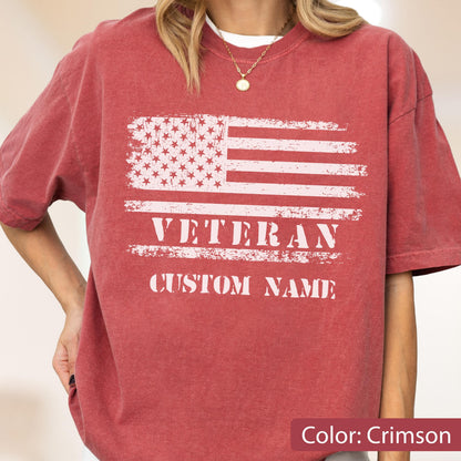 May include: A crimson short-sleeve t-shirt featuring a distressed white graphic of the American flag. Below the flag, the word VETERAN is printed in a bold, distressed white font, followed by the text CUSTOM NAME in the same style.