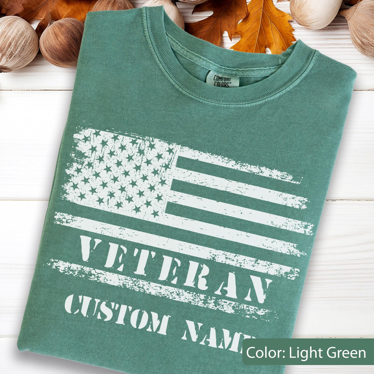 May include: A light green short-sleeved t-shirt featuring a distressed white graphic of the American flag. Below the flag, the text reads VETERAN and CUSTOM NAME in a bold, white, distressed font. The shirt has a Comfort Colors brand label.