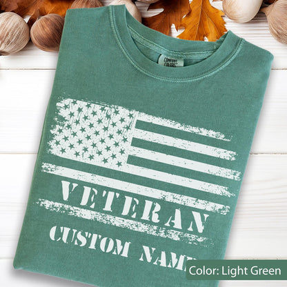 May include: A light green short-sleeved t-shirt featuring a distressed white graphic of the American flag. Below the flag, the text reads VETERAN and CUSTOM NAME in a bold, white, distressed font. The shirt has a Comfort Colors brand label.