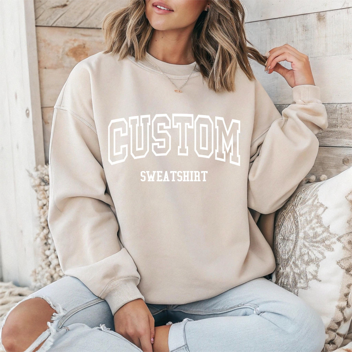 May include: A beige sweatshirt with the word "CUSTOM" in white, block letters above the word "SWEATSHIRT". The sweatshirt is a neutral color and has a relaxed fit. The image shows the sweatshirt on a person.